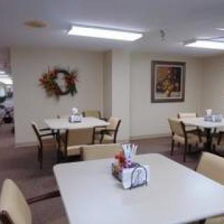 A simple dining area with neatly arranged tables and chairs, featuring wall decor including a wreath and a painting.