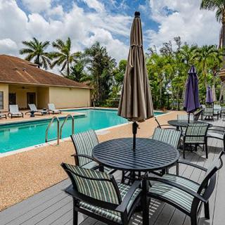 A tranquil outdoor swimming pool area with patio seating and umbrellas, surrounded by lush palm trees.