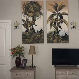 A cozy living room corner featuring tropical wall art, a small television, and a dresser adorned with a lamp and floral arrangement.