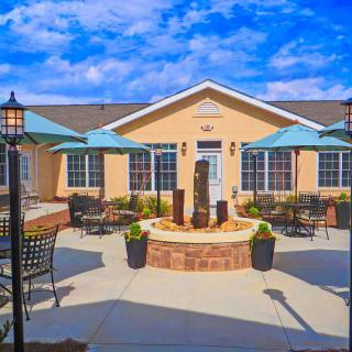A sunny outdoor patio with a central fountain, surrounded by chairs and umbrellas, against a backdrop of a building and blue sky.