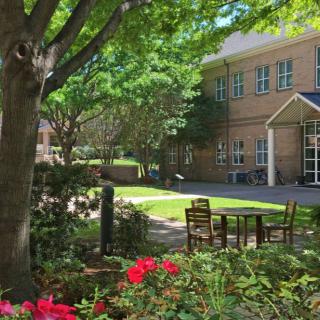 A peaceful garden area with outdoor seating, surrounded by lush trees and vibrant flowers in front of a brick building.