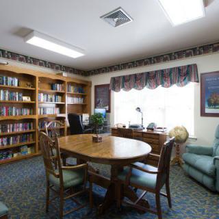 A cozy office space with bookshelves, a round table, and several chairs situated near a large window.