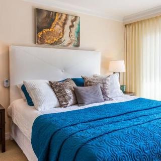A well-decorated bedroom featuring a cozy bed with blue bedding and various pillows, illuminated by natural light and complemented by artwork above the headboard.