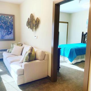 A cozy living room with a white couch and decorative pillows, leading to a bedroom with a bright turquoise bedspread.