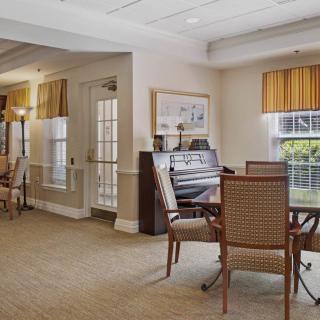 A cozy room with a piano, tables, and chairs, featuring large windows and carpeted flooring.