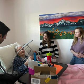 Family visiting an elderly person in a hospital room with a colorful painting on the wall.