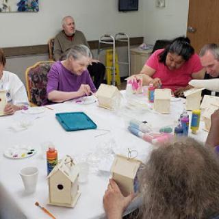 A group of individuals engaged in a creative painting activity, decorating small birdhouses at a community center.