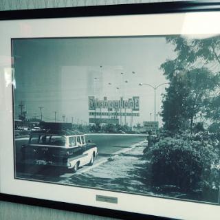 A vintage black and white photograph capturing an urban landscape with a historic car parked on the roadside.