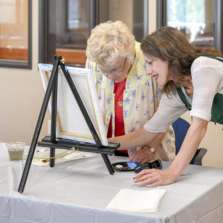 A younger woman assists an elderly woman in a painting class.