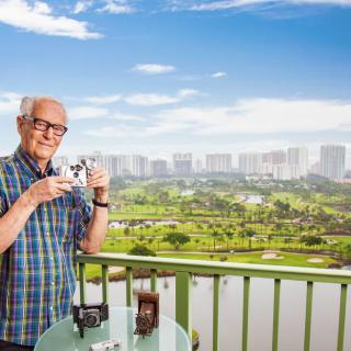 An elderly man stands on a balcony holding a camera, with a city skyline and lush greenery in the background.