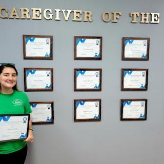 A caregiver proudly holding an award in front of a wall displaying multiple certificates of recognition.