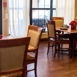 A cozy dining room with wooden tables and chairs, accented by bright natural light streaming through large windows with curtains.
