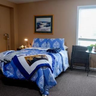 A cozy bedroom with a blue quilt on the bed, a painting on the wall, and a window letting in natural light.