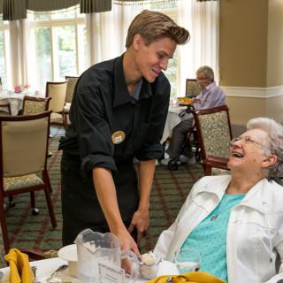 A young adult engages with an elderly person, sharing a moment of laughter in a dining setting.