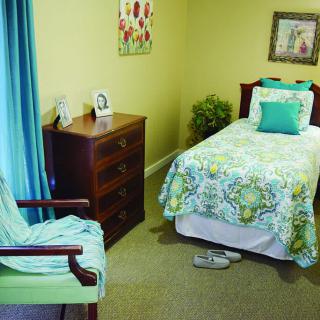 A cozy bedroom with a single bed featuring green bedding and matching curtains, complemented by a wooden dresser and chair.