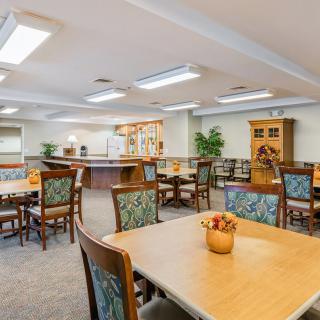 A spacious and well-lit community dining area with multiple tables and chairs, featuring decorative plants and wooden furnishings.