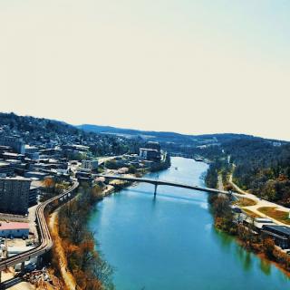 A scenic view of a city alongside a tranquil river, framed by rolling hills under a clear sky.