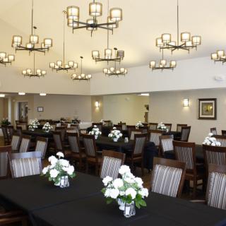 A spacious dining hall with elegant chandeliers, tables set with black tablecloths, and white floral arrangements.