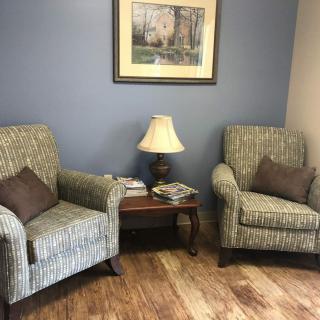 A cozy waiting area with two armchairs, a small table with magazines, and a lamp beneath a framed painting.