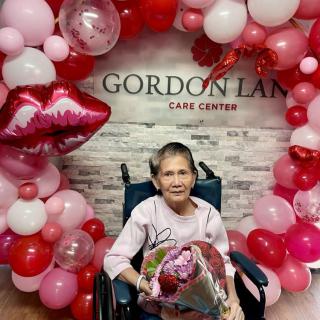 An elderly woman in a wheelchair holding flowers, surrounded by colorful balloons at a care center.