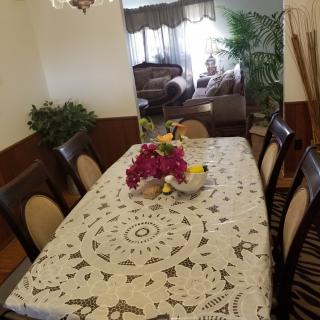 A dining room with a table covered in a decorative lace tablecloth, adorned with a floral centerpiece, and surrounded by cushioned chairs.