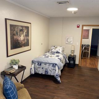 A cozy bedroom featuring a single bed with floral bedding, a side table with a lamp, and a comfortable chair. The room is decorated with a framed painting and has wooden flooring.