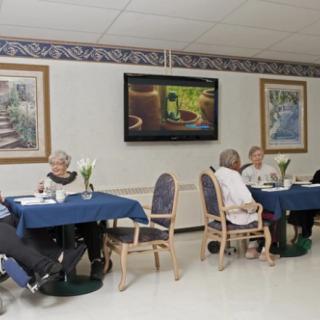 Elderly people sitting at tables in a dining room with a television on the wall.