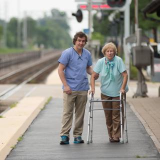 A caregiver assisting an elderly person with a walker at a train station.