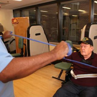A person assists a senior with resistance band exercises in a healthcare facility.