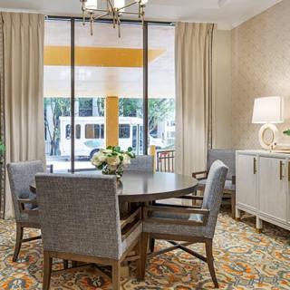A cozy dining area with a round table, four chairs, and large windows adorned with curtains, featuring a stylish lamp and cabinet.