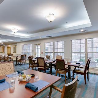 A spacious dining area with neatly arranged tables and chairs, illuminated by ceiling lights and natural light from large windows.