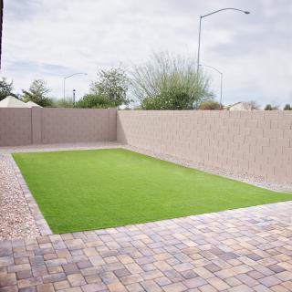 A neatly landscaped backyard with artificial grass and paver walkways enclosed by a tall fence.