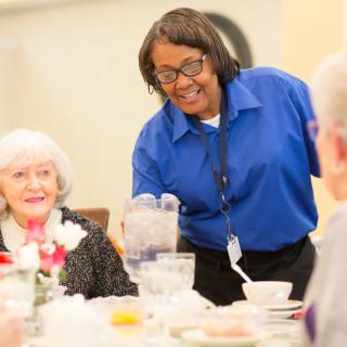 A staff member is serving water to an elderly woman at a dining table.