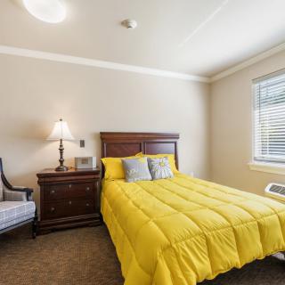A cozy bedroom with a bright yellow comforter on the bed, accompanied by a nightstand with a lamp and an upholstered chair near the window.