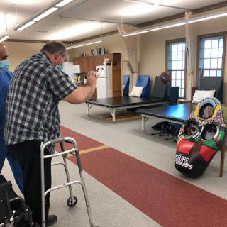 An elderly person using a walking aid participates in a rehabilitation session with the help of a therapist in an indoor setting.