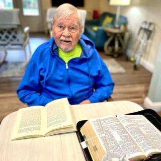 An elderly person sitting at a table with open books, wearing a blue jacket.