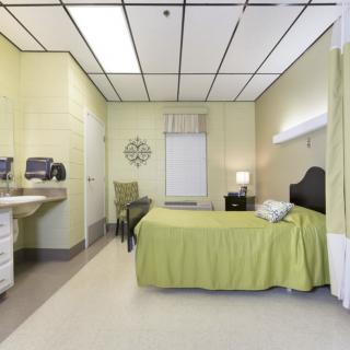 A modern hospital room featuring a single bed with green bedding, equipped with a sink and seating area.