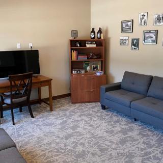 A cozy living room with a sofa, desk and television, featuring a shelving unit and photos on the wall.