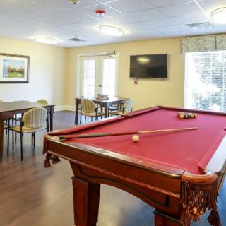 A well-lit game room featuring a red-felt pool table, tables and chairs, and a wall-mounted television.