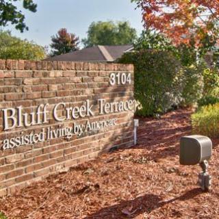 A brick wall sign for Bluff Creek Terrace, surrounded by trees with autumn leaves.