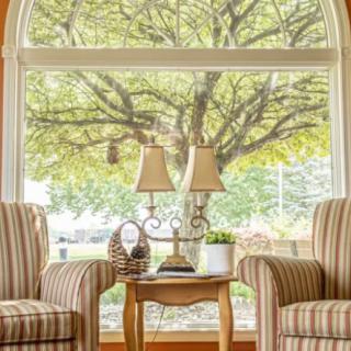 A cozy living room with two striped chairs, a small table with lamps, and a large window showcasing a view of a lush tree.