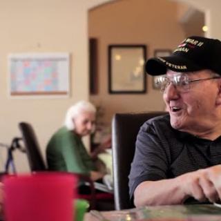 A cheerful elderly man in a hat engages in conversation while another senior woman sits nearby.