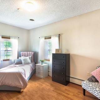 A cozy bedroom with a bed, dresser, and chair, featuring natural light from the windows.