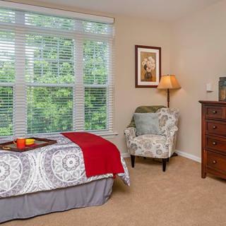 A cozy bedroom with a large window, featuring a bed with a patterned bedspread, an armchair, and a wooden dresser topped with a lamp.