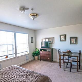 A bright and cozy bedroom with large windows, a bed, a small table with chairs, and a dresser equipped with a TV.