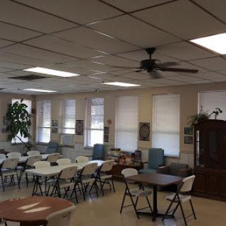 A spacious community room set up with tables and chairs, featuring a ceiling fan and natural light from the windows.
