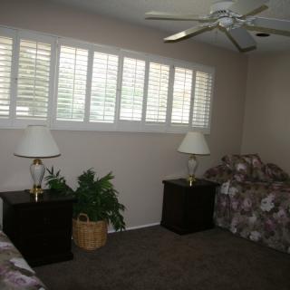 A bedroom interior featuring two twin beds with matching bedding, nightstands with lamps, a ceiling fan, and window shutters.