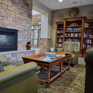 A cozy library corner with comfortable armchairs, a wooden coffee table, and a fireplace set against bookshelves filled with books.