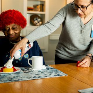 A caregiver adds whipped cream to a dessert for a senior citizen, creating a warm moment of connection.