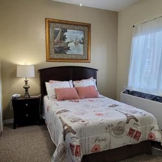 A cozy bedroom with a neatly made bed, featuring decorative pillows and a floral-themed quilt. A lamp sits on a bedside table next to the bed, and artwork is hung above the headboard. Natural light streams in through the window.